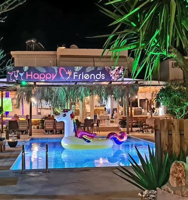 Night view of Happy Friends bar with a unicorn floaty in the swimming pool and outdoor tropical seating.