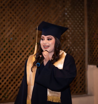 Una mujer graduada sonriente con toga y birrete negros da un discurso frente a un micrófono.