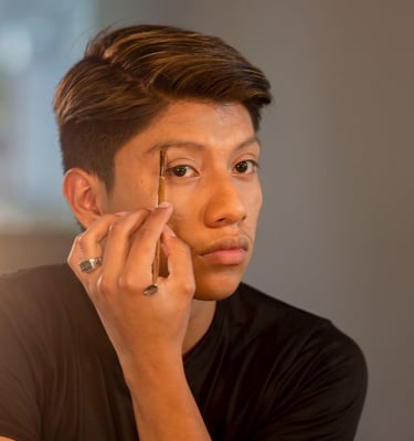 Un joven se aplica maquillaje de cejas con una herramienta de pincel profesional frente a un espejo.