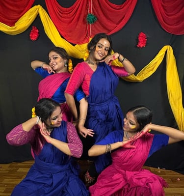 Four Indian women in traditional blue and pink sarees performing a folk dance on a decorated stage.