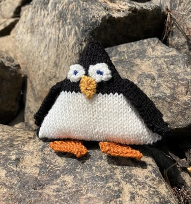 A hand knitted penguin soft toy sitting on rocks