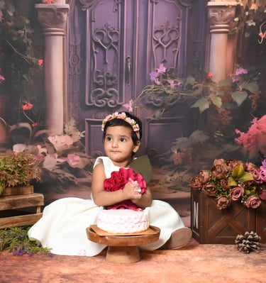 Adorable baby girl in a pink dress, celebrating her first birthday with "ONE" blocks, balloons, and floral backdrop.