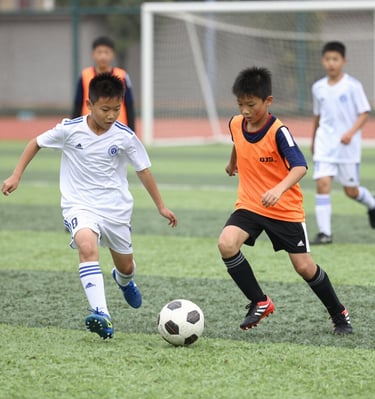 A group of young people, some dressed in sports uniforms, are gathered together. The scene appears to be at a sports event, possibly a soccer match, with several individuals wearing shirts labeled 'Barça Academy.' One person in a bright orange top stands out, engaging in conversation with another in a bright red and yellow outfit. Referees in green uniforms are also present in the background.