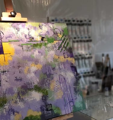 A purple, yellow, and green abstract painting on an easel with paints in the background