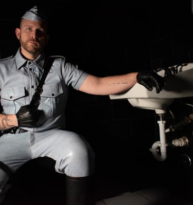 man dressed in leather squatting in low light beside sink