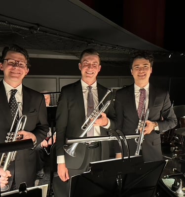 Three men holding trumpets and wearing suits, in an orchestra pit.