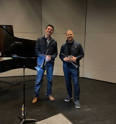 George Chase and Caleb Hudson smile, holding piccolo trumpets, during a rehearsal break.