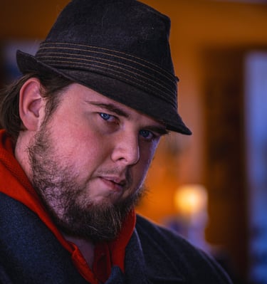 A man with a beard wearing a dark fedora and jacket in a close-up portrait in soft lighting. Athens,