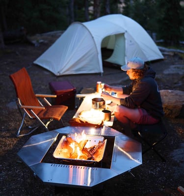 a man sitting at a table with a plate of food next to a campfire