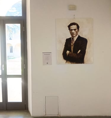 Portrait painting of Pier Paolo Pasolini displayed on a white art gallery wall next to a glass door.
