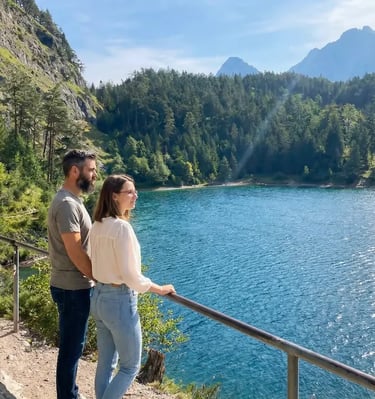 Couple standing at viewpoint railing overlooking turquoise Drachensee lake surrounded by pine forest
