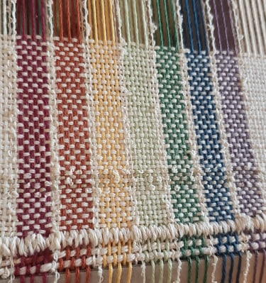 a loom with an unfinished hand-woven towel in muted rainbow pride flag colours