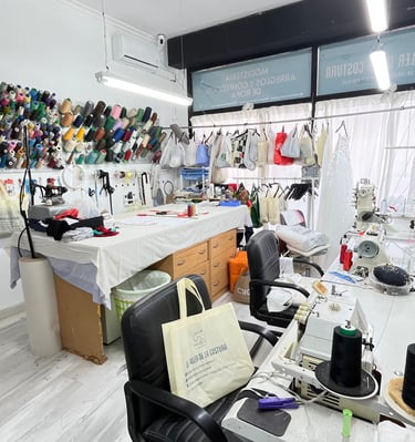 Vista general de un taller de costura con varias máquinas de coser, mesa de corte, rollos de tela, h