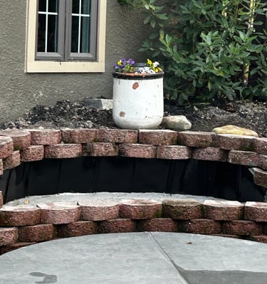 Curved fire pit retaining wall made of red stone blocks next to a white flower pot and house window.