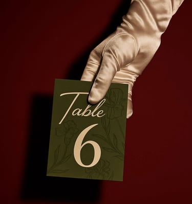 A hand in a satin glove holds an elegant green wedding table number card against a red background.