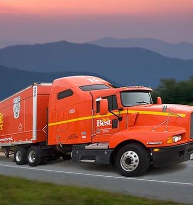 An orange Do it Best semi-truck driving on a mountain highway at sunset.