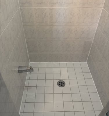 A walk-in shower featuring white floor tiles, a chrome drain, marble-patterned wall tiles, and a bathtub spout.