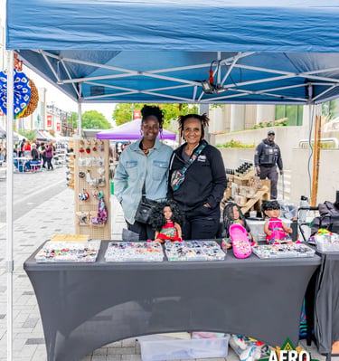 Two smiling women standing at their craft stall under a blue canopy at the Afro Fiesta Festival.