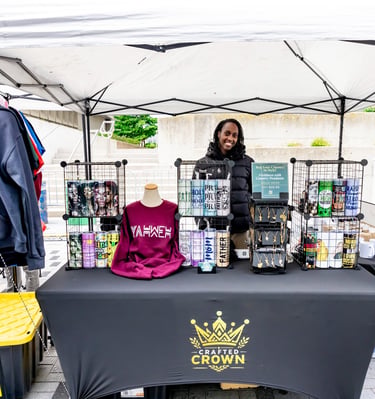A smiling vendor showcases custom apparel and printed tumblers at the Crafted Crown booth during the Afro Fiesta Festival.