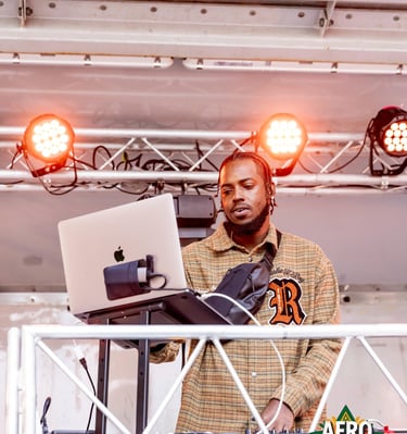 A male DJ performing a live music set with a laptop and mixer under orange stage lights at Afro Fiesta Festival.