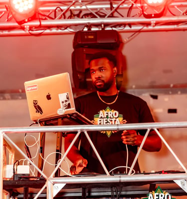 A DJ performing at the Afro Fiesta Festival under red stage lights with a laptop and turntable.