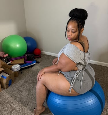 A curvy woman in grey loungewear sits on a blue yoga ball during a home workout session.