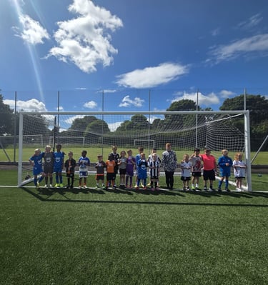 A youth football team posing together in front of a goal on a sunny outdoor astro pitch.