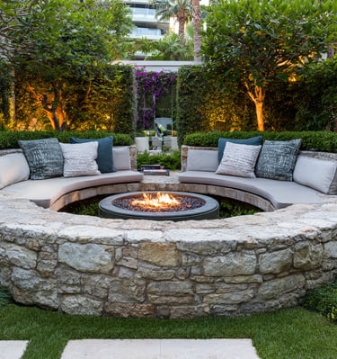 Luxury circular stone fire pit with built-in curved seating and cushions in a landscaped garden patio.