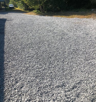 A wide gray crushed stone gravel driveway leading past green trees to a parked car.