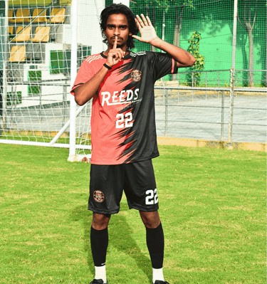 A soccer player in a red and black jersey making a silence gesture on a green football pitch.