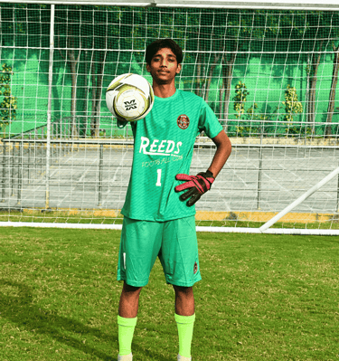 A youth football goalkeeper in a green kit holding a soccer ball in front of a goal post on a grass pitch.