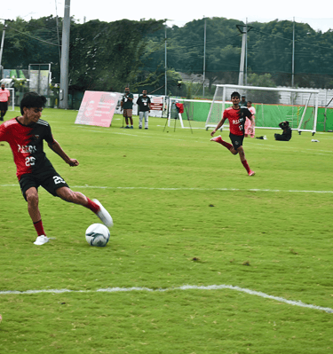 A soccer player in a red and black jersey dribbles a soccer ball on a green field during a match.