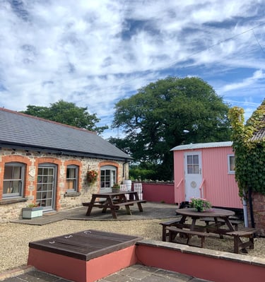 Rustic stone cottage and pink shepherd hut with outdoor wooden picnic tables on a sunny gravel patio.
