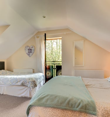 A bright attic bedroom with two comfortable beds and a balcony overlooking green trees.
