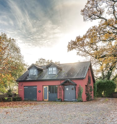 Red country cottage with grey doors and gravel driveway surrounded by autumn trees under a bright sky.
