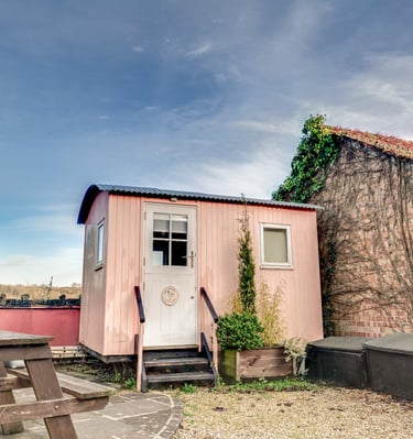 A small pink shepherd's hut with a white door next to a rustic brick outbuilding.