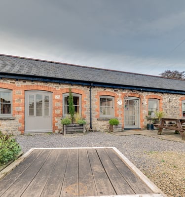 Rustic stone holiday cottage with grey doors, gravel courtyard, and wooden picnic table.