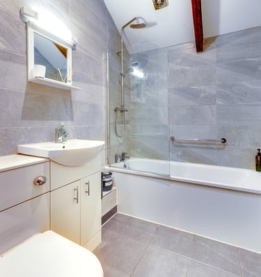Modern bathroom with grey stone tiles, a white bathtub with a glass shower screen, and a vanity sink.