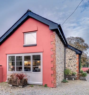 A charming pink stone cottage with a gravel driveway and outdoor seating area under a blue sky.