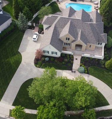 Aerial view of a luxury suburban home with a gray shingle roof, large lawn, and swimming pool.