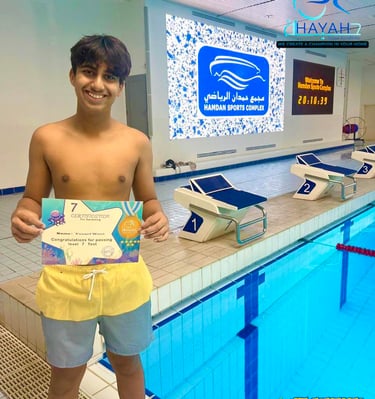 A young male swimmer holding his Level 7 swimming certificate at Hamdan Sports Complex pool.