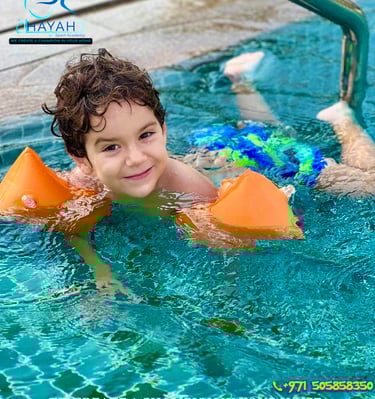 A smiling young boy with orange floaties learns to swim in a pool during Hayah Sport Academy lessons.