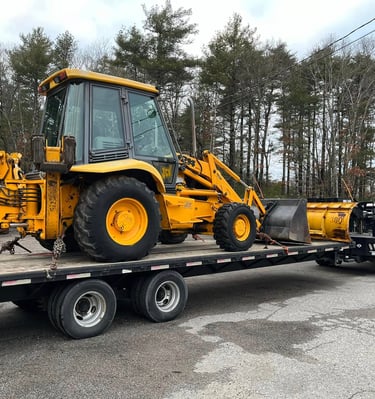 Yellow JCB backhoe loader on a flatbed trailer with a plow, ready for transport.