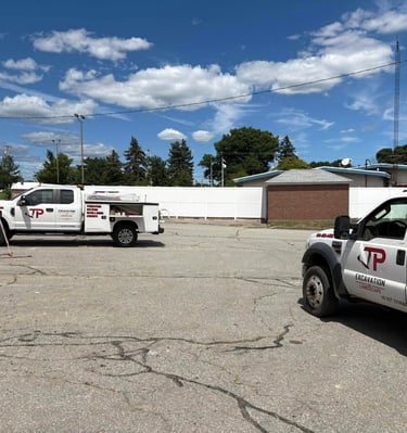 Two JP Excavation & Landscape trucks, a white service truck and a dark dump truck, parked on asphalt under a blue sky, ready