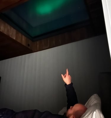 A person lying in bed points up at the glowing green Northern Lights visible through a glass ceiling skylight.