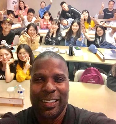 Dr. Marlon Ware standing in front of his class with students holding up the peace sign
