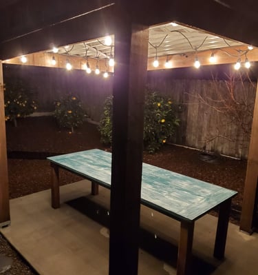 Reclaimed wood outdoor dining table under custom pergola with metal roof and lights. Colorado Springs remodeling.