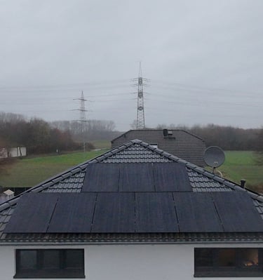 Residential rooftop with black solar panels installed on dark tiles overlooking a landscape with power lines.