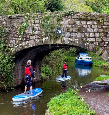 A family paddleboard under a stone bridge on the Monmouthshire and Brecon canal in the Brecon Beacons, surrounded by nature.