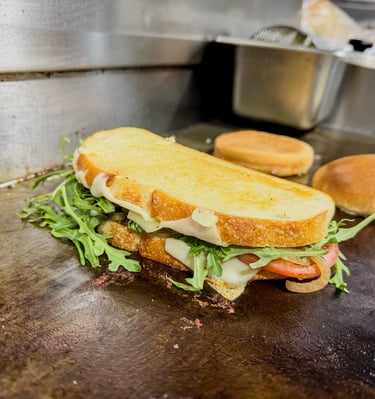 A gourmet panini with melted cheese, fresh arugula, and tomato on a flat top grill.